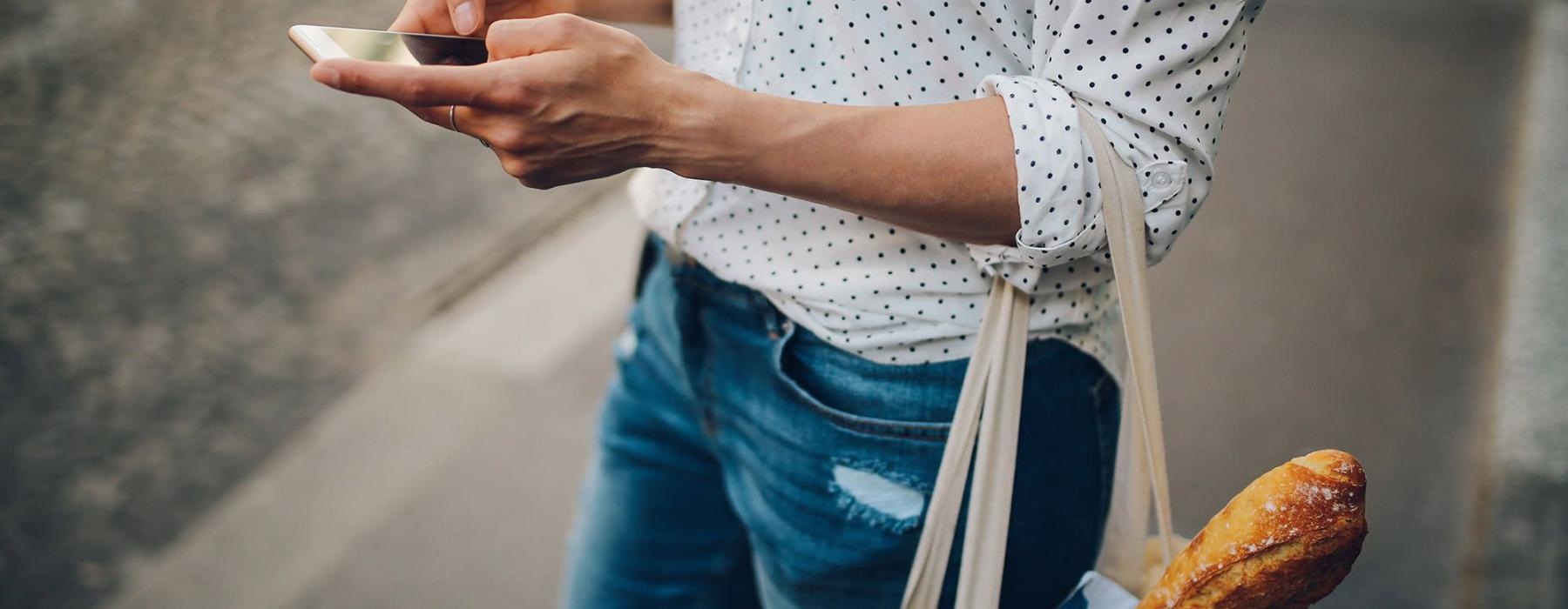 woman walks down the street and texts with a bag of groceries on her arm